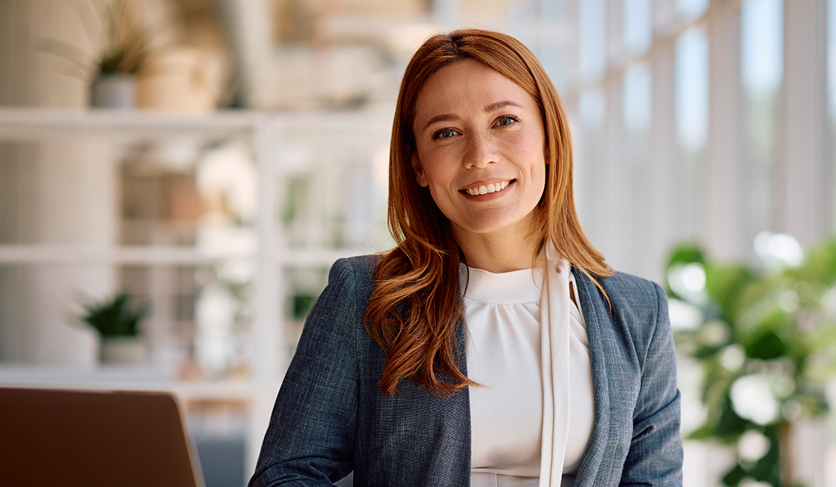 A smiling woman with red hair in a gray blazer sits at a desk in a bright modern office