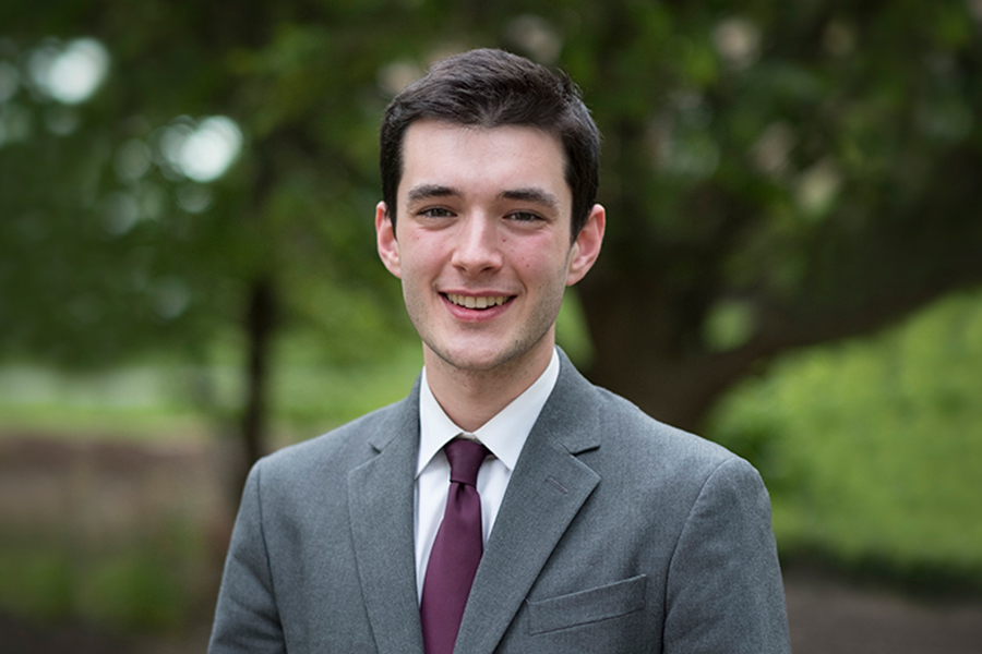 A professional portrait of a smiling young man with dark hair, wearing a grey suit jacket, white dress shirt, and a purple tie. He is standing outdoors with a soft-focus background of green trees.