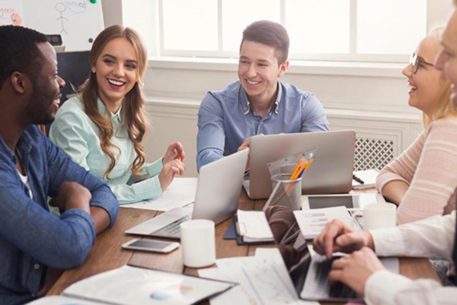 A diverse group of five young professionals smiling and collaborating around a sunlit conference table with laptops and notebooks.