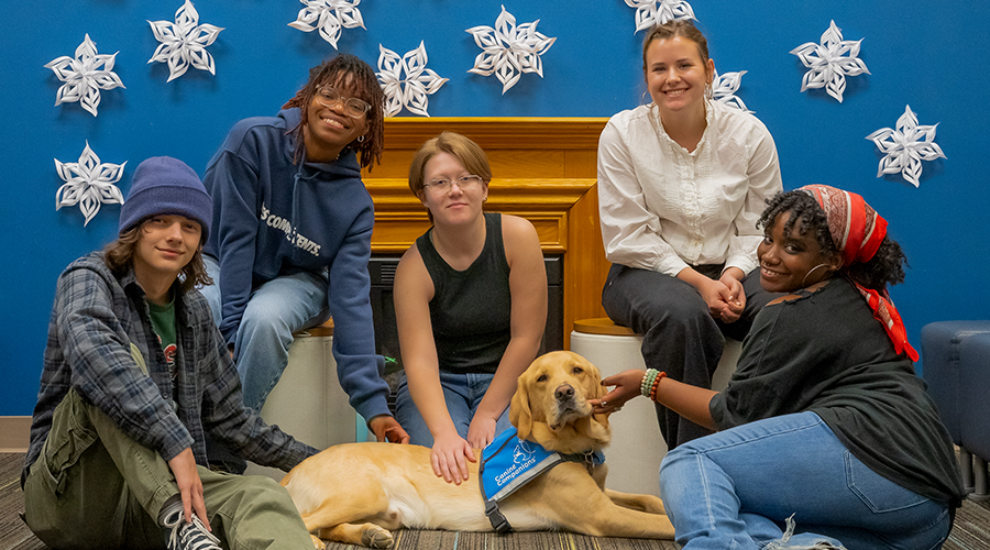 Five smiling young adults posing with a yellow Labrador Retriever wearing a blue 